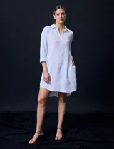 Vestido camisero blanco de algodón con cuello solapa, escote en V y mangas 3/4 regulables. Presenta bolsillos laterales verticales y abertura con botones en la espalda.