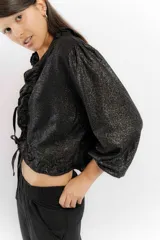 Blusa corta negra con textura brillante, cuello con volados y mangas abullonadas 3/4. Se ata en la parte delantera con cordones.
