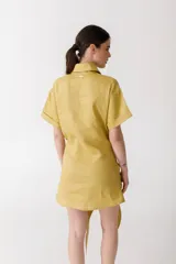 Vestido corto color pistacho, estilo camisa, con cuello clásico, mangas cortas con dobladillo y cierre frontal parcial con botones. Presenta un lazo anudado en la cintura.