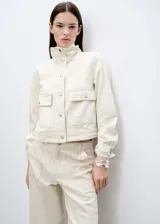 Chaqueta corta color crema, de estilo tejano vaquero, con cuello alto, cierre frontal con botones metálicos, mangas largas con puños abotonados y dos bolsillos de parche con solapa y botón en la parte delantera.