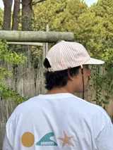 Gorro de visera curva con diseño a rayas verticales en tonos rosado pálido y blanco. La corona tiene un bordado en azul claro con texto. La visera es de color beige claro. Incluye un cordón negro ajustable para colgar del cuello.