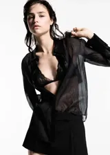 Camisa negra semitransparente de tejido fluido con ribetes satinados, cuello camisero, cierre de botones y mangas largas abotonadas.