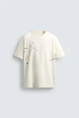 Camiseta color crudo de corte holgado, cuello redondo y manga corta. Tiene estampado de salpicaduras de pintura negra en el frente y texto estampado en la espalda.