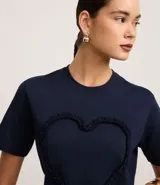 Remera azul marino de algodón con cuello redondo y mangas cortas. Presenta un aplique en forma de corazón con volados en el frente.