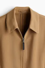 Campera color marrón claro de tejido suave con cuello clásico, cierre frontal con cremallera, bolsillos laterales insertados al bies y puños ajustables con botón a presión.