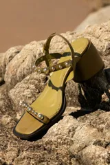 Sandalias de cuero color verde oliva con taco medio y punta cuadrada. Tiras con tachas esféricas plateadas y cierre con hebilla en el tobillo.