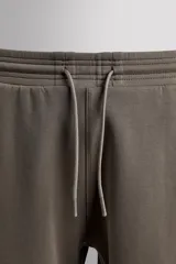 Pantalón jogger color marrón oscuro, corte regular fit, confeccionado en tejido técnico compacto de algodón con elasticidad. Cuenta con cintura elástica ajustable con cordón, bolsillos frontales y bolsillo lateral con cierre de cremallera.