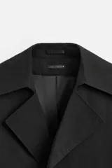 Trench corto negro confeccionado en tejido técnico ligero, con cuello con solapas de muesca, manga larga y cierre frontal de botonadura cruzada.
