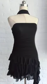 Vestido negro, sin mangas, con escote asimétrico y detalle de lazo o corbatín largo que cae desde el hombro. El cuerpo es ajustado y fruncido, y la falda corta tiene volados superpuestos.