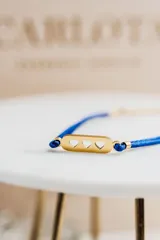 Pulsera con hilo azul brillante y detalles dorados en acero quirúrgico. Dije rectangular dorado con tres corazones calados.
