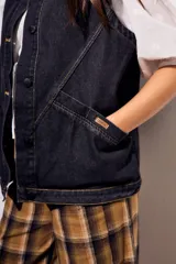 Chaleco oversize de denim negro con costuras en hilo ocre, cuello camisero, cierre frontal con botones y bolsillos delanteros.