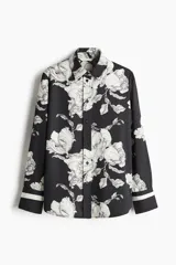 Camisa de sarga estampada con flores en blanco y negro, de corte recto, con cuello clásico, cierre frontal con botones, mangas largas con puños abotonados y bajo redondeado.