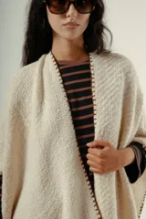 Ruana tejida a mano en lana merino 100%, de color marrón con detalles de costura en rojo en los bordes y terminación de flecos largos en el ruedo. Presenta una silueta amplia y caída natural, confeccionada artesanalmente por Manos del Uruguay.