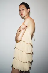Minivestido color crema con cuello halter ajustable con lazadas y espalda descubierta. Presenta volantes en el mismo tejido.