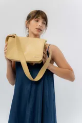 Cartera tipo bandolera de eco cuero texturado color amarillo pálido, con solapa frontal que cierra con imán oculto y correa regulable. Presenta el logo metálico de la marca en la parte inferior de la solapa.