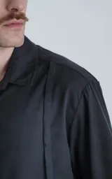 Camisa negra de manga corta con cuello clásico y cierre frontal con botones. Presenta una tira vertical cosida en el frente.