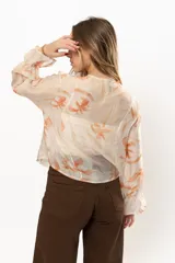 Camisa color crema con estampado floral en tonos naranja, con cuello en V y volados frontales. Mangas largas abullonadas con puños elásticos.