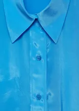 Camisa celeste de tejido satinado, con cuello camisero, cierre frontal asimétrico con botones, manga larga con puños abotonados, diseño ajustable con cuerda en la parte posterior y corte largo.