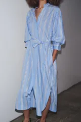 Vestido midi de algodón a rayas celestes y blancas con cinturón de la misma tela, cuello redondo, manga abullonada con puño y cierre frontal con botones.