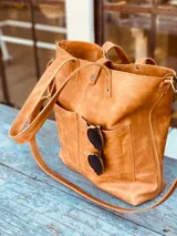 Cartera tote de cuero color tostado, con doble asa de mano y correa larga extraíble y ajustable. Tiene cierre superior y dos bolsillos externos. Interior forrado con compartimento acolchado para laptop de hasta 14 o 16 pulgadas.