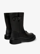 Bota de media caña color negro, con diseño de suela gruesa y estriada. Presenta una correa con hebilla en la parte superior de la caña y un logo de la marca "Camper" en una etiqueta.