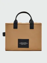 Bolso tote mediano de lona color beige con detalles en negro. Tiene doble asa de mano de cuero y correa bandolera ajustable y extraíble. Cuenta con dos bolsillos frontales con solapa y un bolsillo frontal con cierre. Estampado frontal con la leyenda "THE TOTE BAG" y el logo "MARC JACOBS".