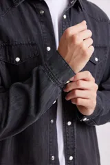 Camisa de jean de manga larga, color gris oscuro lavado, con cuello y botones a presión blancos. Presenta dos bolsillos frontales con solapa y pespuntes visibles.