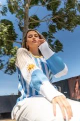 Sweater tejido a rayas verticales celestes y blancas, con cuello redondo amarillo y detalle bordado del sol de la bandera uruguaya en la manga.