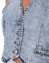 Chaleco corto de denim celeste con tachas plateadas, cuello en V y cierre frontal con botones.