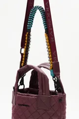 Cartera tipo tote mini acolchada, color negro brillante, con asas cortas y correa larga ajustable. La correa larga es intercambiable y presenta un diseño trenzado multicolor con tonos turquesa, amarillo y negro.