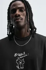 Camiseta negra de corte relaxed fit, cuello redondo y manga corta. Estampado frontal con dibujo de gato y texto en japonés.