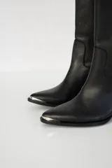 Botas bucaneras de cuero negro con puntera fina y taco cuadrado bajo. Presentan una tira de cuero vertical con apliques metálicos y hebilla decorativa en la parte superior de la caña.