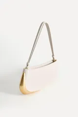 Bolso de hombro alargado color crema, con asa corta y detalle metálico dorado en un lateral. Tiene un bolsillo interior con cremallera y cierre principal con cremallera.