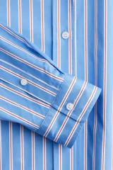 Camisa oversized de popelina de algodón a rayas verticales celestes, blancas y rojas. Tiene cuello clásico, cierre frontal con botones, hombros caídos y mangas largas con puños abotonados. El bajo es redondeado.