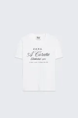 Camiseta blanca de algodón con cuello redondo y manga corta. Estampado frontal con el logo de Zara, la inscripción "A Coruña" en letra cursiva y la leyenda "Established 1975 A Rúa Juan Flórez 64-66".