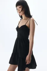 Vestido corto negro de popelina de algodón con breteles finos ajustables con moña, escote corazón y cuerpo entallado con costuras decorativas y espalda fruncida. Falda acampanada con costura fruncida en el bajo.