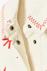 Sobrecamisa color crudo de Zara Woman Collection, con cuello solapa y manga larga. Tiene bolsillos de parche en el delantero y bordados combinados a contraste en color rojo. Cierre frontal con botones a presión ocultos.