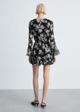 Vestido corto negro con estampado floral blanco, de mangas largas acampanadas, cuello redondo, lazo en la cintura y volados en el ruedo.