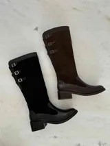 Bota alta de cuero negro con caña de gamuza negra y detalles de hebillas plateadas. Tiene un taco cuadrado bajo y cierre lateral. El diseño combina texturas y detalles metálicos para un estilo moderno y elegante.