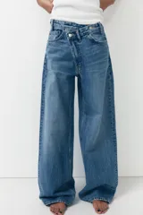 Pantalón de jean celeste de tiro alto, corte wide leg y largo hasta el tobillo. Presenta un diseño asimétrico en la cintura con cierre frontal cruzado con cremallera y botones metálicos.