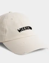 Gorra de gabardina color crudo con visera curva y detalle de roturas. Bordado frontal con la palabra "Weekend" en negro.