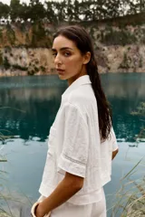 Camisa blanca de cuello solapa y manga corta, con bolsillo delantero y detalle de vainicas a tono. Cierre frontal con botones ocultos por solapa.