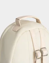 Mochila color crema de tela acolchada con pespuntes verticales. Tiene un bolsillo frontal con cierre, doble cierre superior, manija superior, correas regulables y bolsillos laterales. Incluye un charm colgante de corazón.