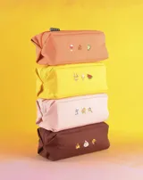 Neceser naranja de tela con cierre superior y logo de la marca en etiqueta negra. Presenta bordados de emojis: copa de vino, fuegos artificiales y mano verde.