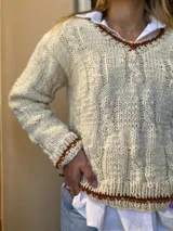 Sweater artesanal de lana color crudo con diseño de trenzas y ochos. Tiene cuello redondo y terminaciones en marrón.