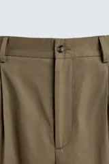 Pantalón color caqui de corte tapered relaxed fit, confeccionado en tejido de algodón. Presenta cintura con pliegues frontales dobles, bolsillos laterales en cadera y bolsillos traseros de vivo con botón. Cierre frontal con cremallera y botón.