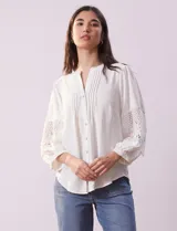 Blusa blanca de gasa con cuello a la base, escote en V y abertura central con botones. Tiene mangas 3/4 amplias con apliques de crochet y detalles plisados en la parte delantera.