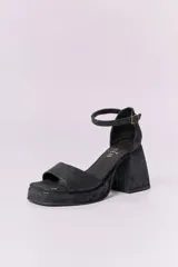 Sandalias de cuero negro con plataforma y tacón cuadrado alto. Presentan una tira delgada sobre los dedos y pulsera tobillera con hebilla dorada.
