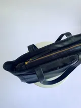 Cartera tipo tote de cuero graneado color negro, con textura uniforme y suave al tacto. Tiene manijas de 55 cm, bolsillo frontal con broche imantado, bolsillo trasero con cierre metálico, cierre principal metálico, forro interior y tres bolsillos internos (uno con cierre, uno plaqué y uno tipo sobre con solapa y botón).