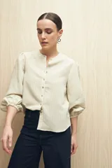 Camisa color crema a rayas verticales finas, de cuello mao, mangas 3/4 abullonadas con elástico en los puños y cierre frontal con botones de nácar.